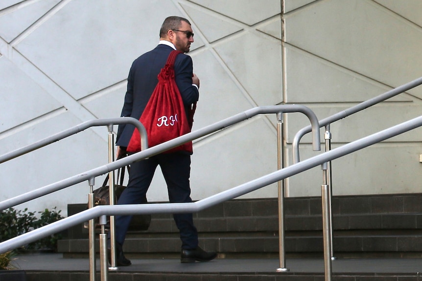 A man wearing a suit and sunglasses carrying a red sack and a small bag walks towards the entry to the District Court in Perth.