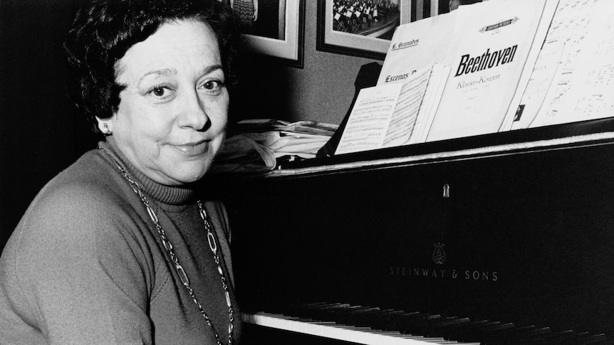 A black and white photograph of Spanish pianist Alicia de Larrocha sitting at the piano, smiling with her hands folded.