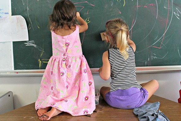 Children sit on a table while using chalk to draw on a school blackboard.