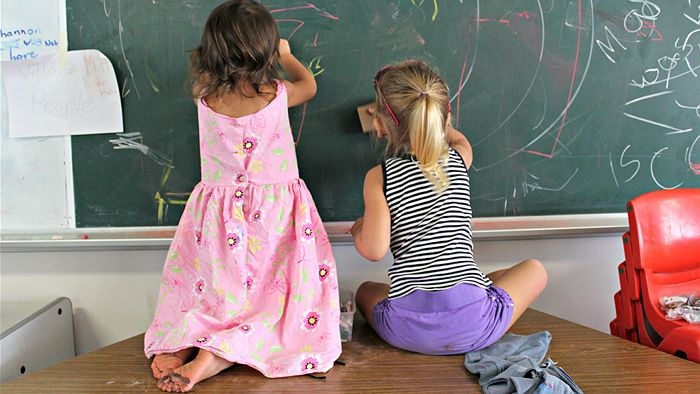 Children sit on a table while using chalk to draw on a school blackboard.