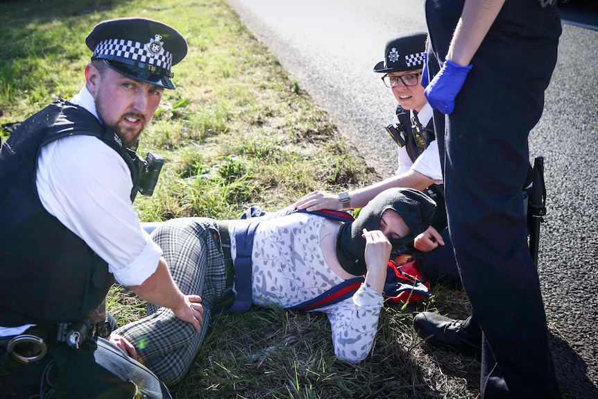 Two police officers crouch down, holding their hands to a woman in a balaclava who has been pushed to the ground
