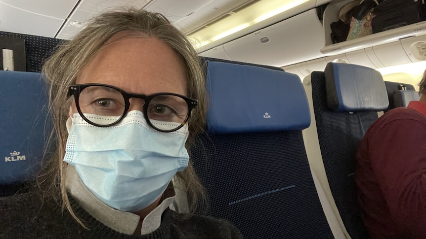 A woman wearing black rimmed glasses and a disposable face mask sits in her seat on a plane