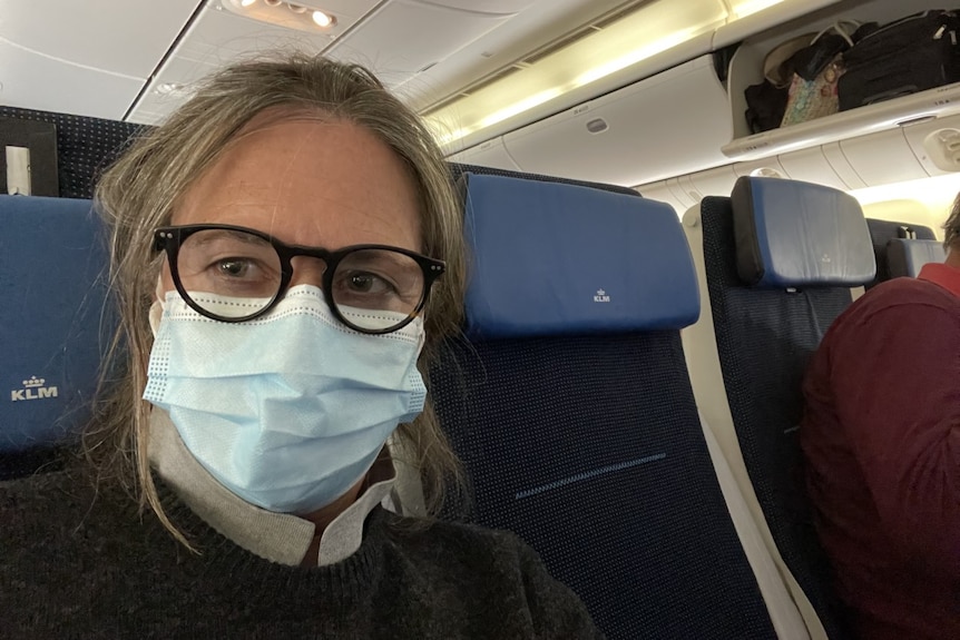 A woman wearing black rimmed glasses and a disposable face mask sits in her seat on a plane