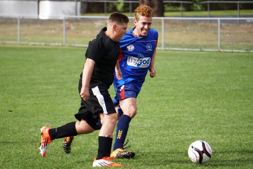 Two young men run behind a soccer ball on a field, one in a bright blue uniform, the other in black with a white stripe.
