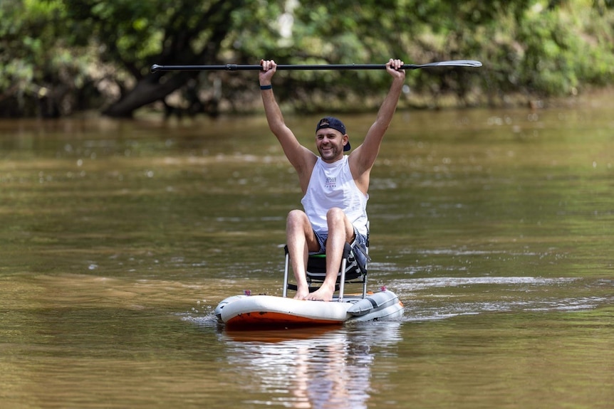 A man in white singlet sitting on an inflatable paddle board on flat waters, his arms raising a paddle