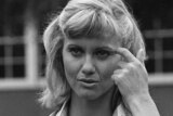 A black and white photo of Olivia Newton John fixing her hair and holding a book, playing Sandy in Grease