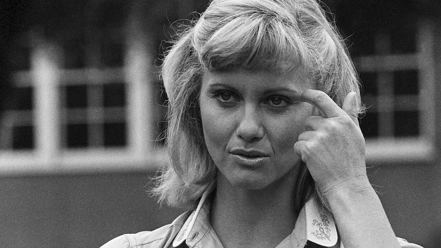 A black and white photo of Olivia Newton John fixing her hair and holding a book, playing Sandy in Grease