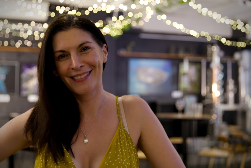 A smiling woman with long dark hair standing with twinkling lights behind her