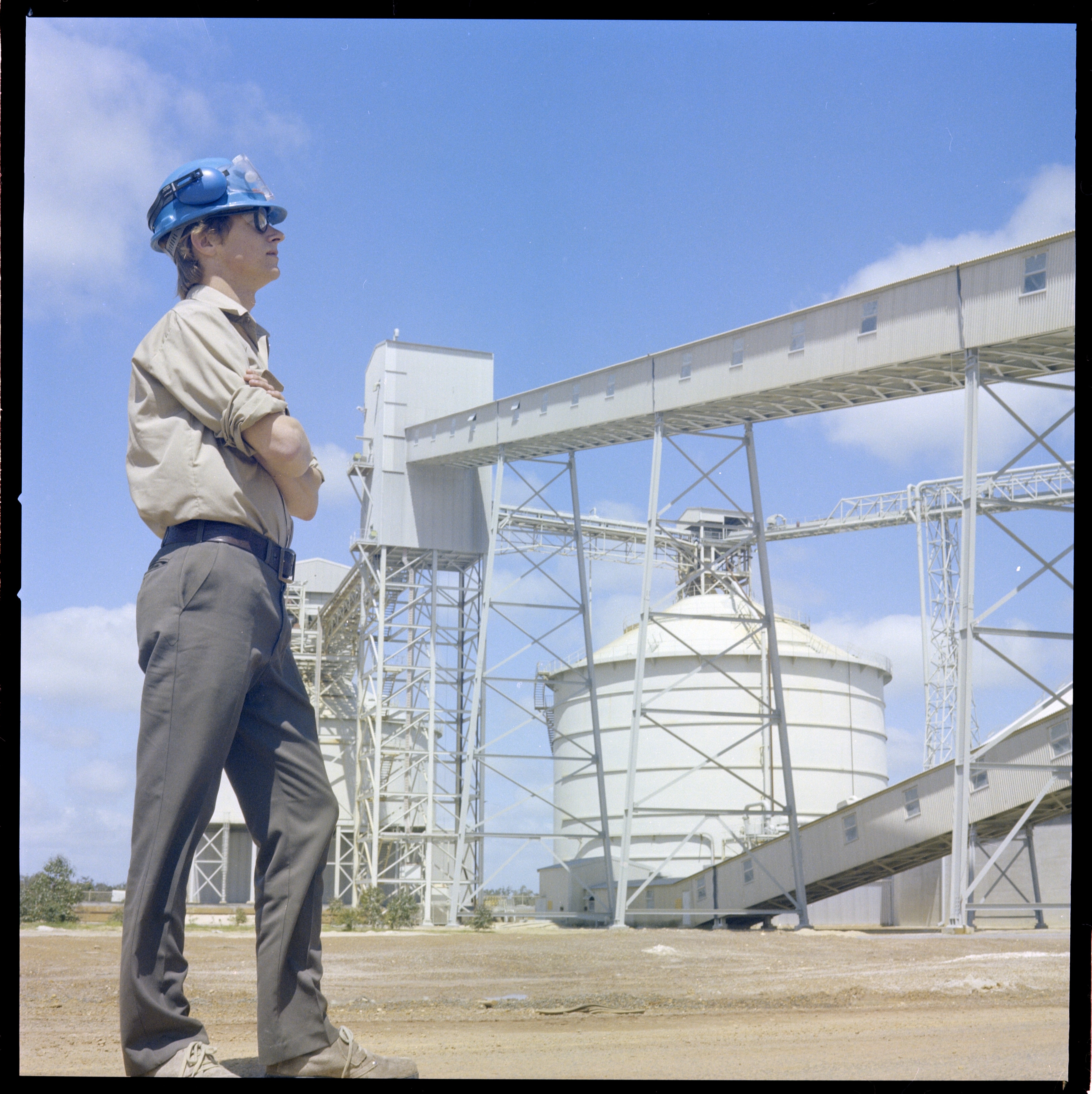 A man in a hard hat crossing his arms, looking at a bauxite refinery