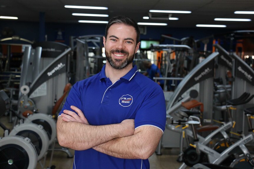 Mr Elliot smiles with his arms folded. He stands in front of workout machines.