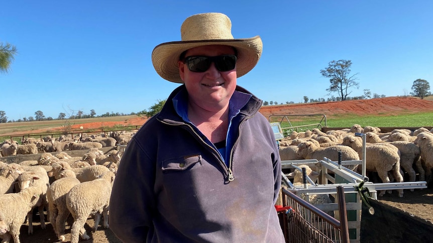 A farmer in sunglasses and a wide hat with sheep in the background