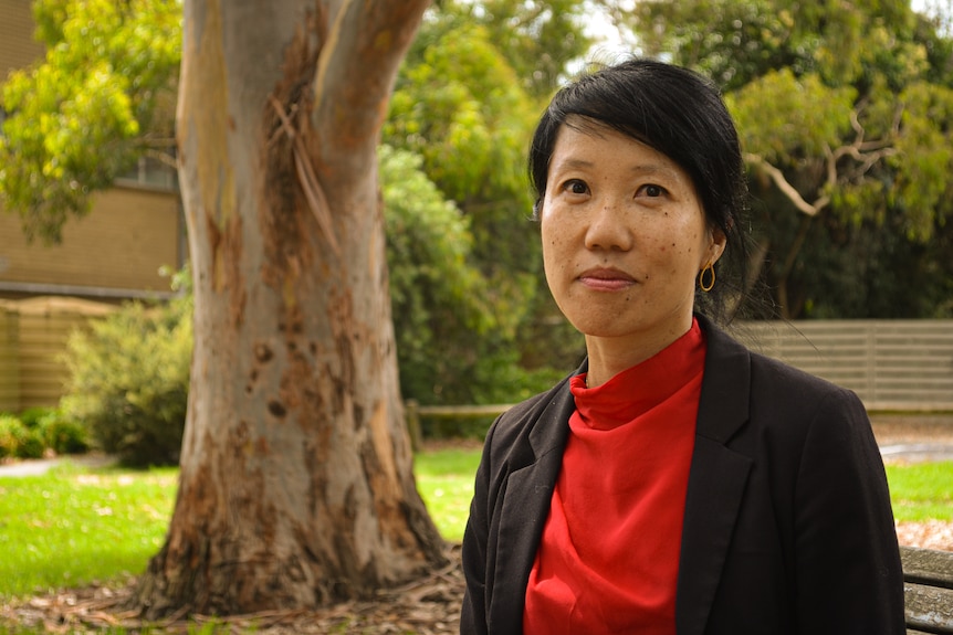 Belinda Lo wearing a red top and a black blazer standing outside infront of a large gum tree