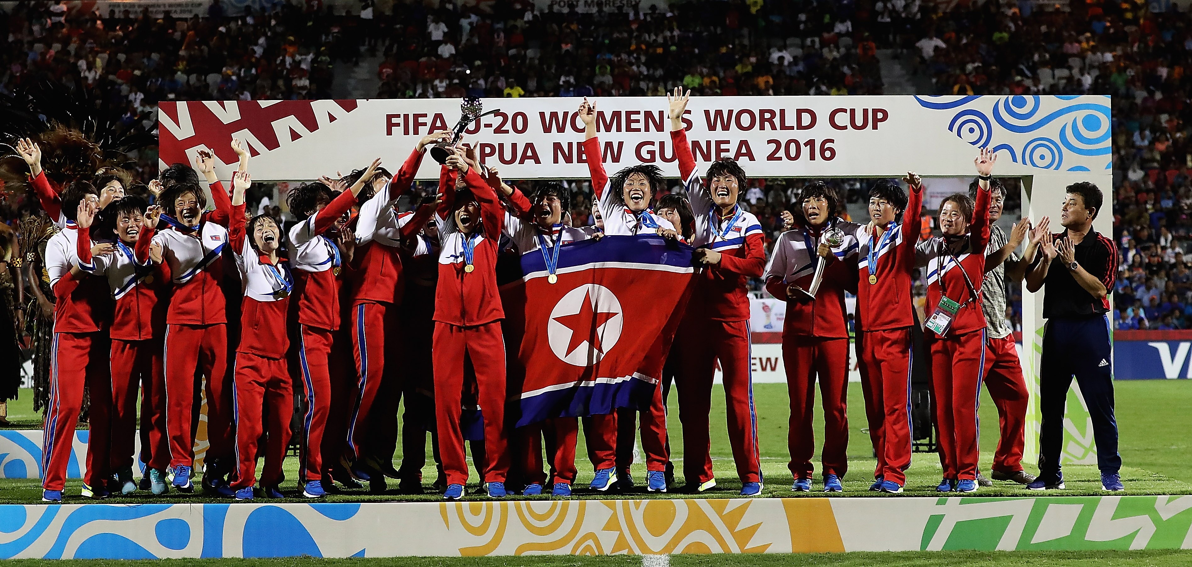 A soccer team wearing red, white and blue tracksuits celebrates winning a tournament on stage holding a flag