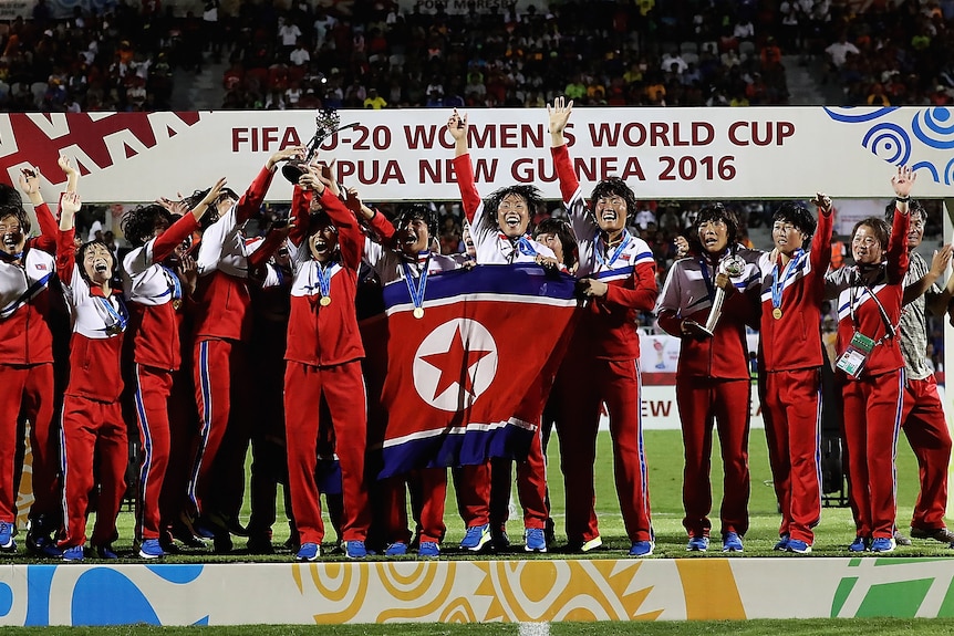 A soccer team wearing red, white and blue tracksuits celebrates winning a tournament on stage holding a flag