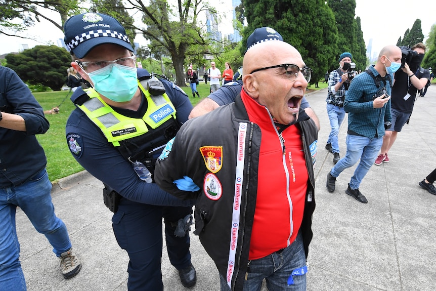 A police officer detains a man in  a red shirt who is not wearing a mask.