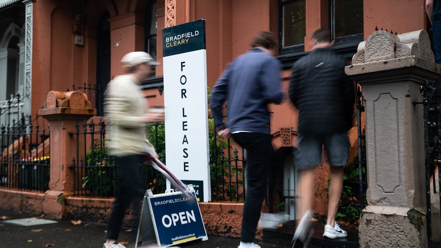 People walk through the front door of a house, past a For Lease sign.