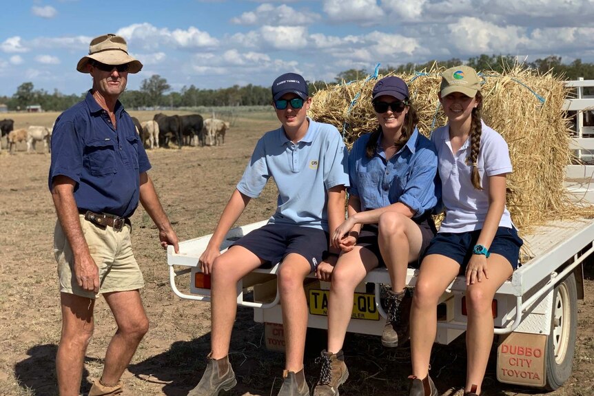 A farmer stands to the left of his three teenage children sitting on the back of ute loaded with hay