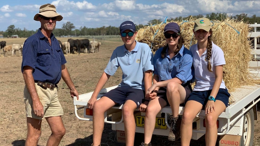 A farmer stands to the left of his three teenage children sitting on the back of ute loaded with hay