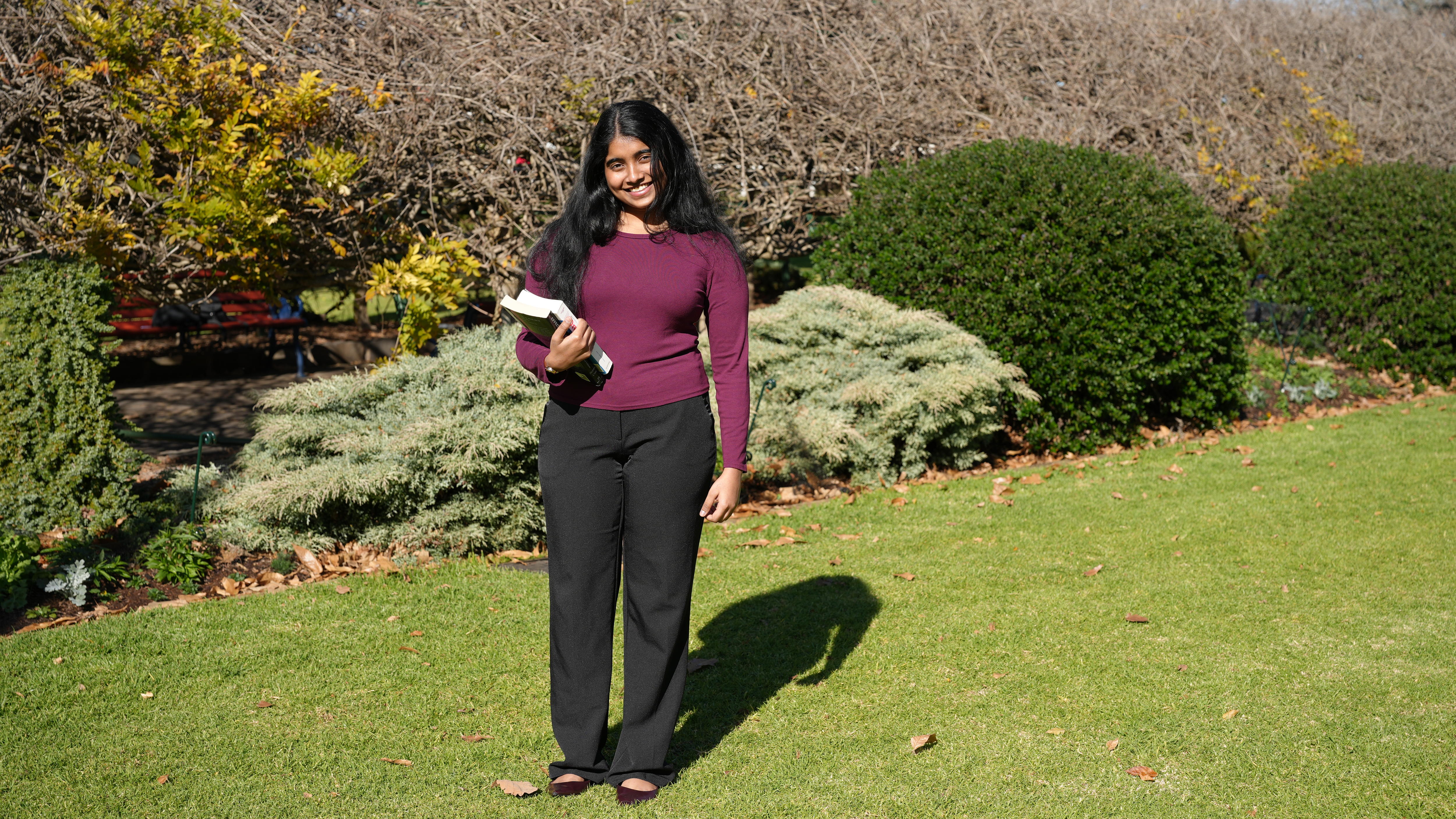 A woman wearing a burgundy top and black trousers smiles in a garden setting. 