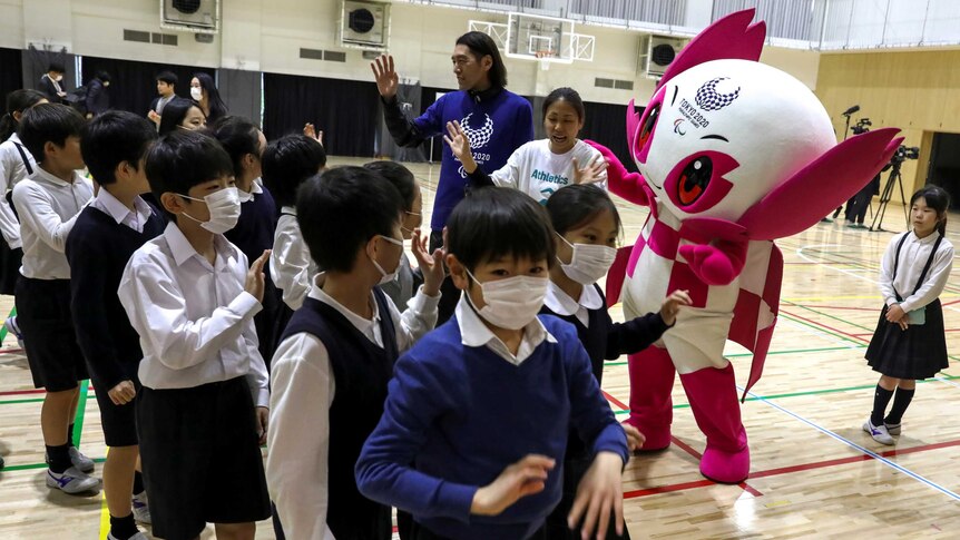 Young students wear masks as they greet a Japanese Olympic mascot.