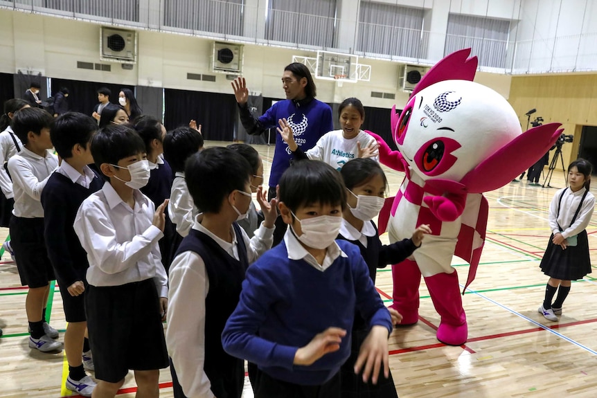 Young students wear masks as they greet a Japanese Olympic mascot.