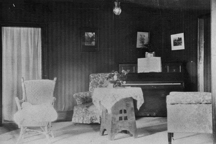 A black and white photo of a basically furnished living room with an old upright piano.