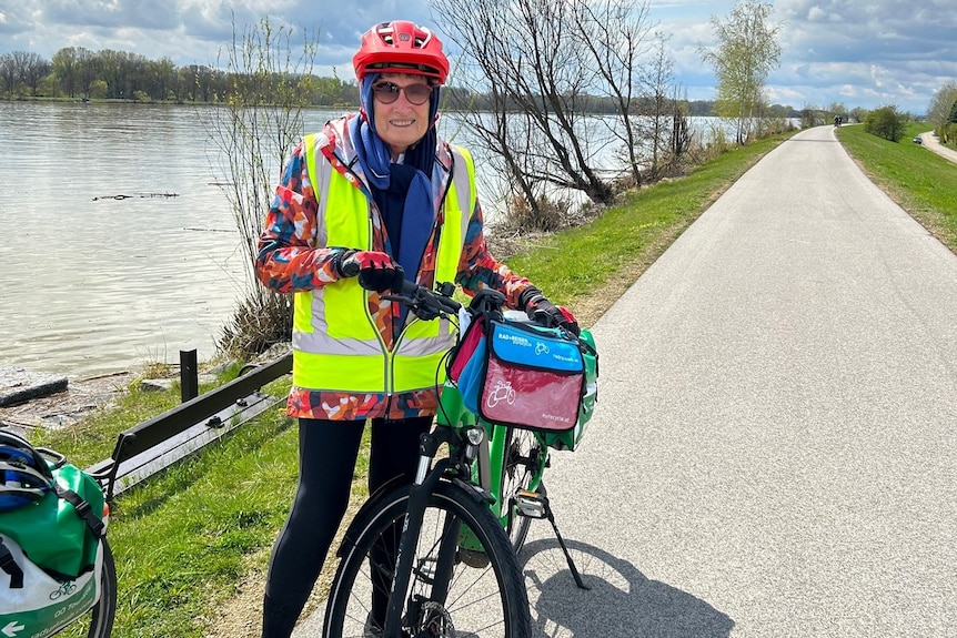 A woman in a fluro vest, scarf and red helmet holds a bike next to a river