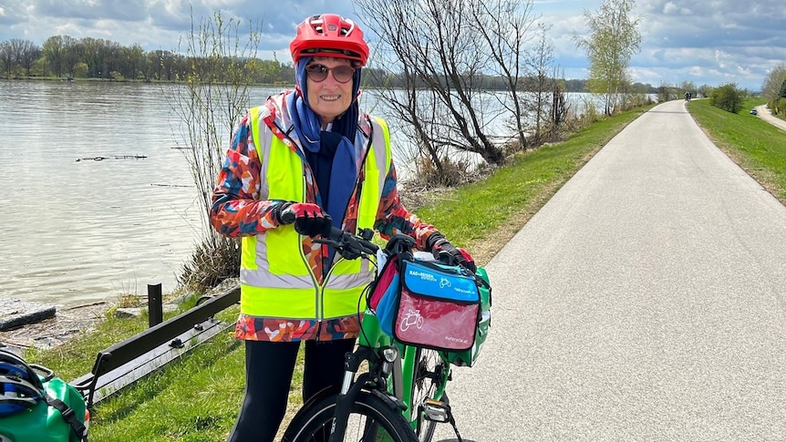 An older woman in a fluro green vest, scarf and red helmet holds a bike next to a river