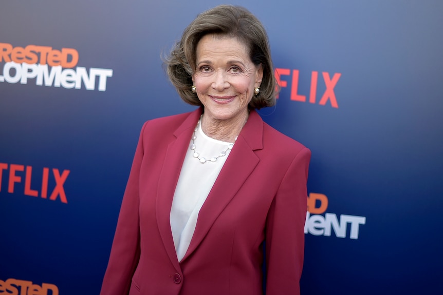 A woman with short brown hair is smiling and wearing a pink blazer.