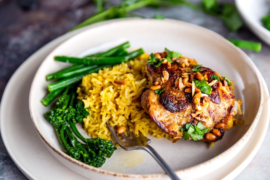 Plate of saffron rice, baked chicken thigh topped with roasted nuts and parsley, served with broccolini.
