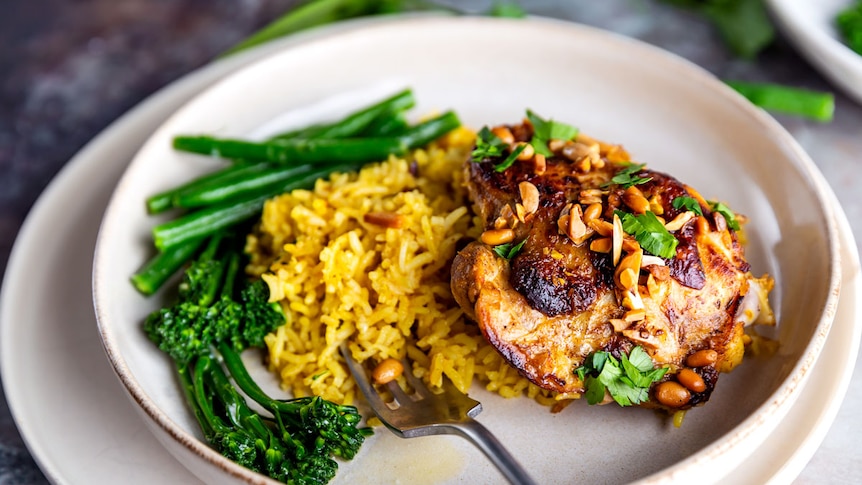 Plate of saffron rice, baked chicken thigh topped with roasted nuts and parsley, served with broccolini.