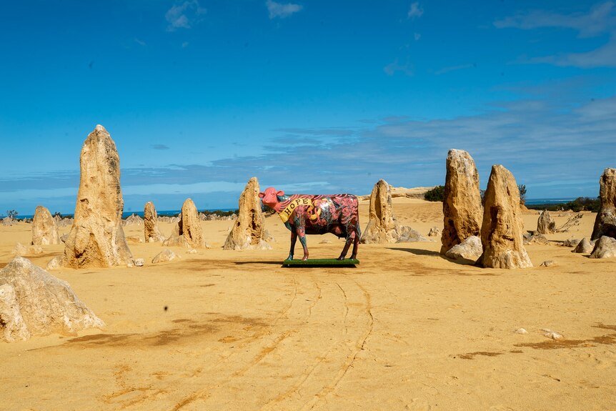 A pink cow sculpture is the distance stands on sand among protruding limestone rocks. 