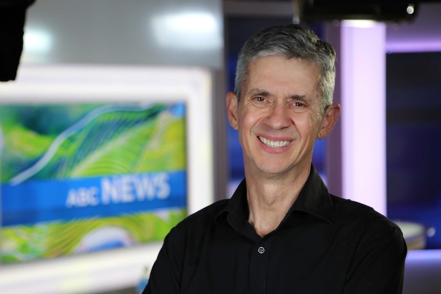 A smiling Eric Napper standing in front of ABC News logo on TV screen in news studio.