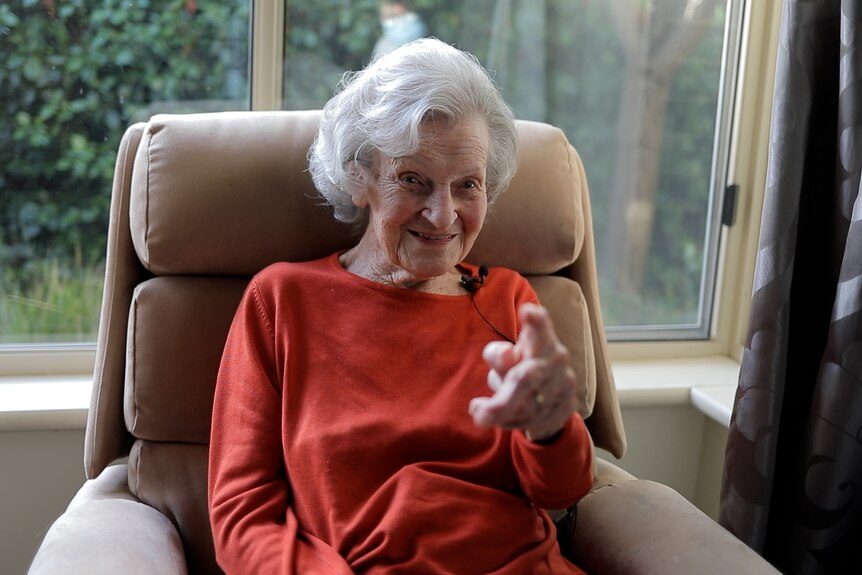 An elderly woman sits in a chair in a red shirt, pointing at the camera.