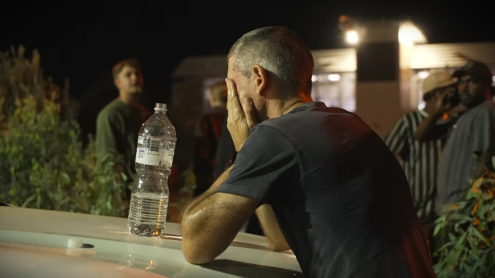 Un hombre blanco se apoyó contra el capó de un automóvil, con una gran botella de agua llena sobre el capó, las manos sobre los ojos y la cara, y se inclinó hacia adelante.