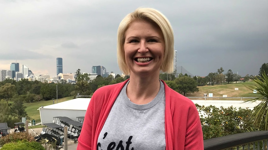 A blonde woman wearing a grey jumper and salmon cardigan smiles at the camera, outside on a gloomy day.