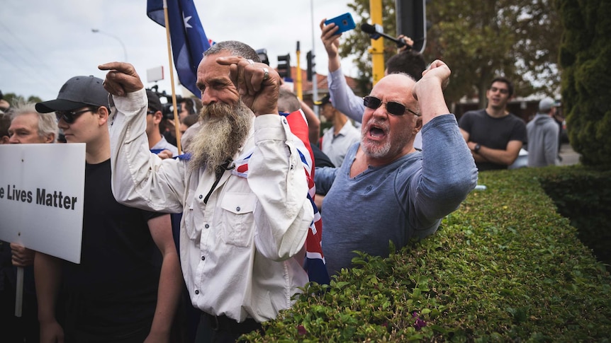 A group of protesters stand shouting and pointing fingers while holding placards and Australian flags outside.