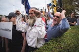 A group of protesters stand shouting and pointing fingers while holding placards and Australian flags outside.