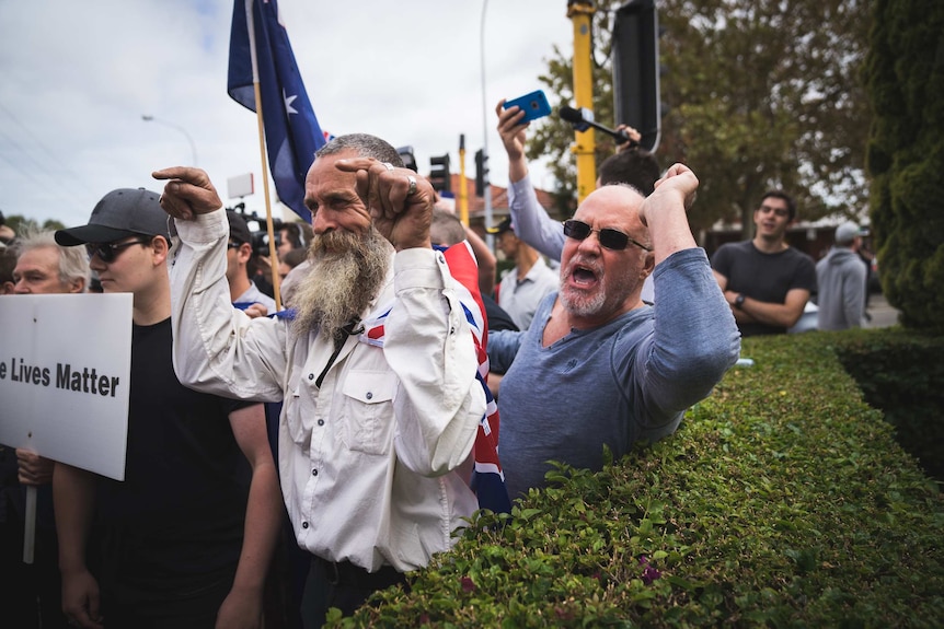 A group of protesters stand shouting and pointing fingers while holding placards and Australian flags outside.