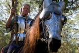 A black horse with an armour headpiece in the foreground, with a man in shiny armour sitting on the horse.
