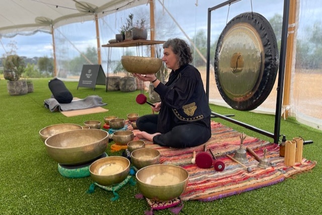 A woman sits in front of a gong, with metal bowls in front of her.