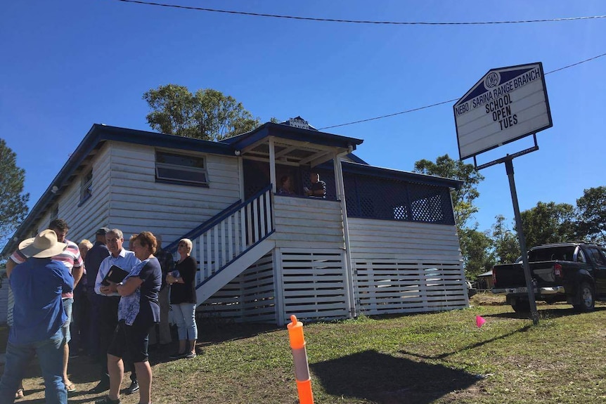 People stand in front of a Queenslander hall and chat. A sign outside the hall says 'school open Tues'