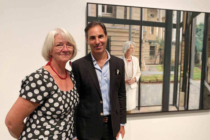 A man and a woman stand in front of a portrait of the woman. 