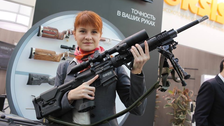 Maria Butina, a young woman with red hair, poses with a large gun, smirking at the camera.