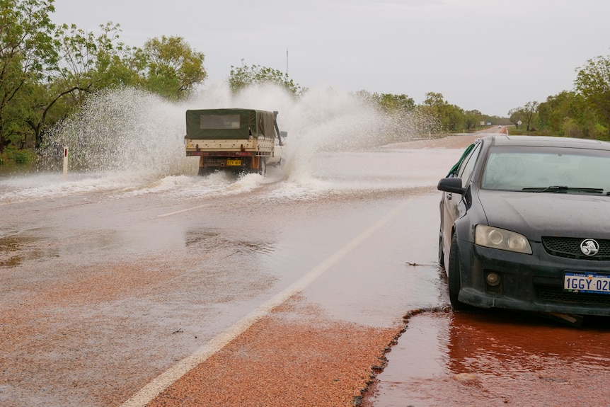 A ute causes a splash as it drives through water on a highway, with a dark sedan pulled over on the side of the road
