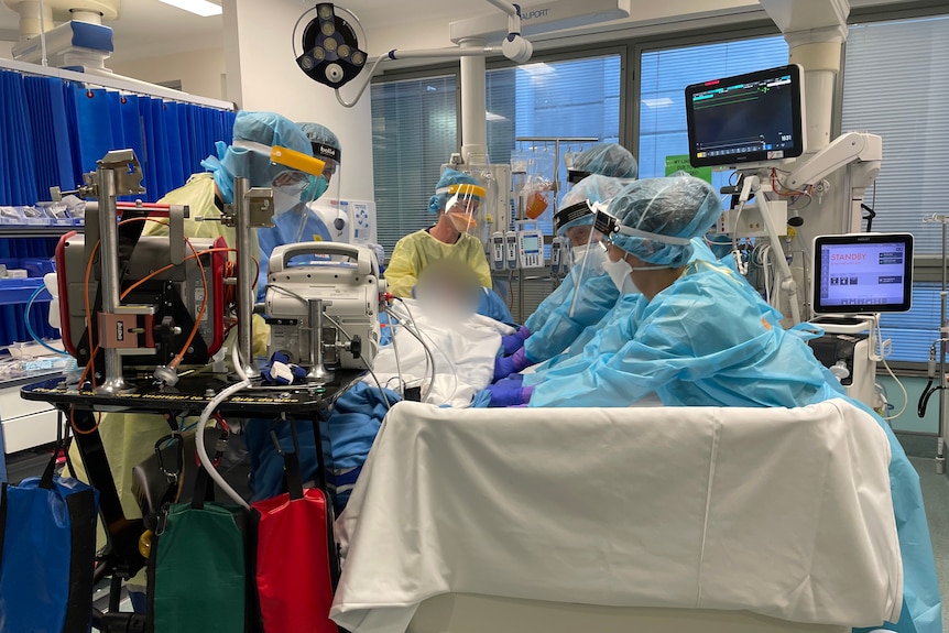 Six people wearing PPE move a patient in an ICU bed.