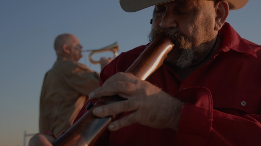 A man plays the digeridoo while another man plays the trumpet in the background