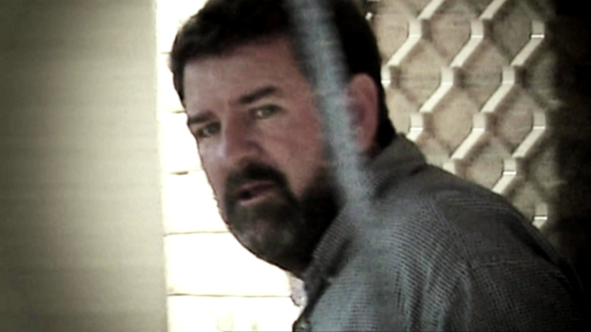 A man with short dark hair and a beard looks sideways at the camera