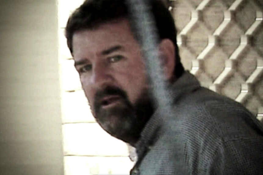 A man with short dark hair and a beard looks sideways at the camera