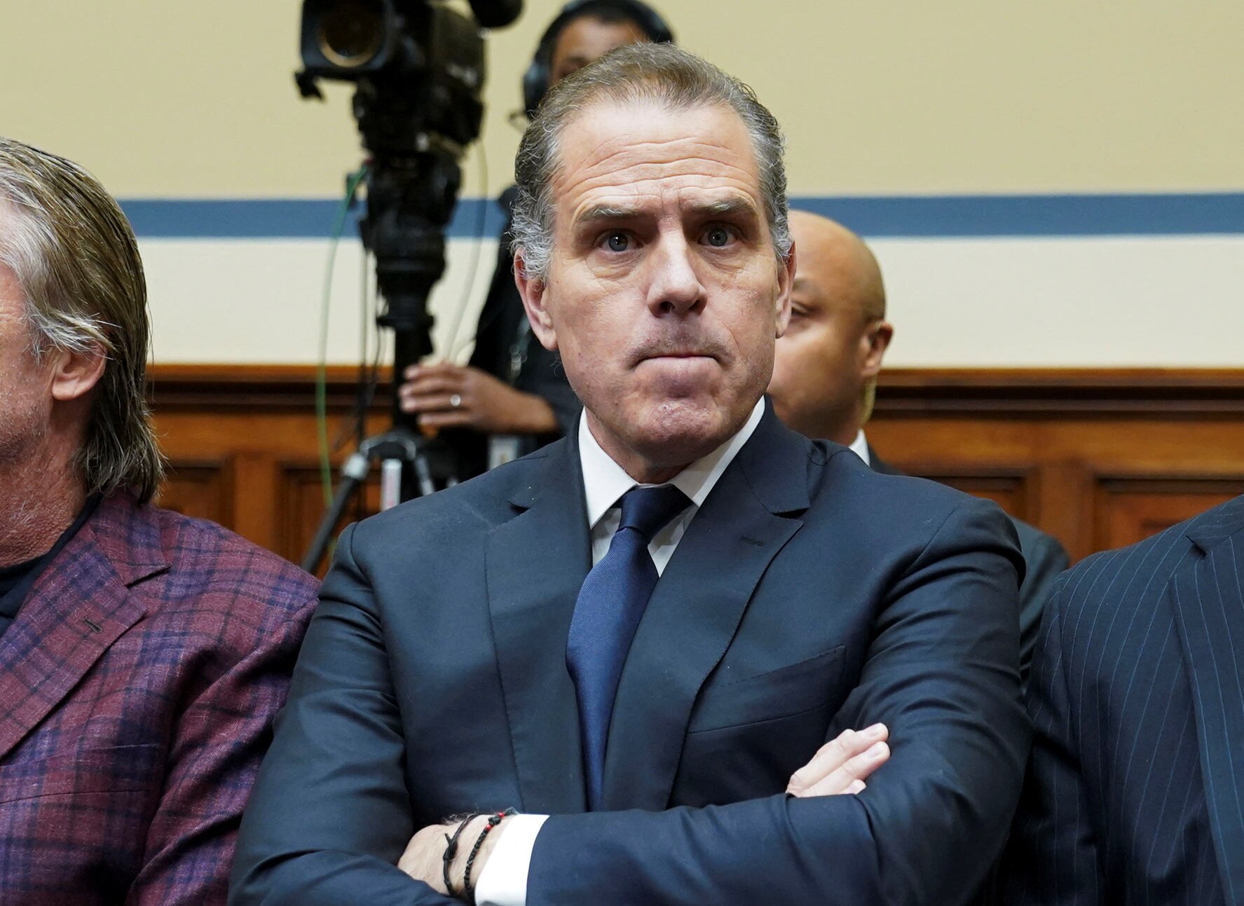 A close-up of Hunter Biden, sitting between people in a Committee room, wearing a suit and tie and crossing his arms.
